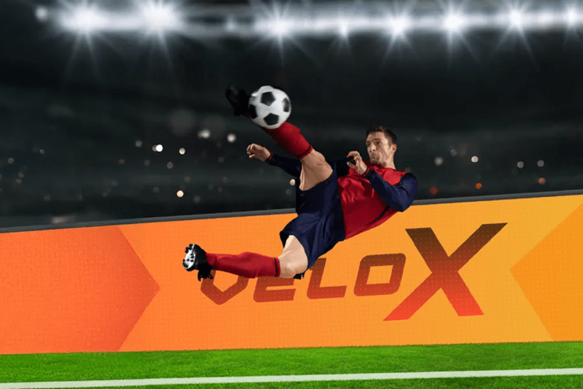 Soccer player in mid-air kicking a ball in a brightly lit stadium with an orange "veloX" ad board.