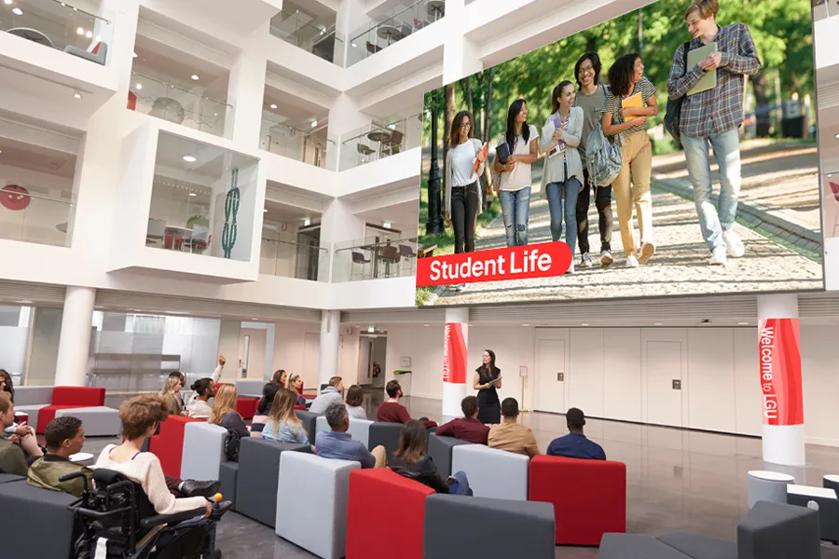University students attend an orientation in a student commons. A large LG DVLED video wall displays a video about student life.