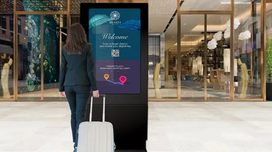 A woman in business dress pulling a wheeled luggage case behind her. She's paused to look at a map displayed on an outdoor digital display.
