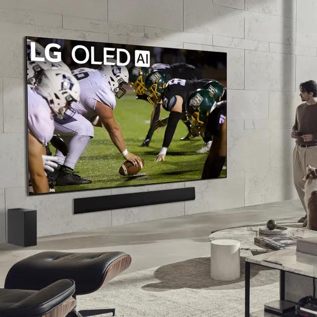 In a cozy, comfortable and well-lit modern living room setting, a man with a dog enjoys a American football game on an LG OLED TV featuring AI technology, immersed in the excitement of the match.