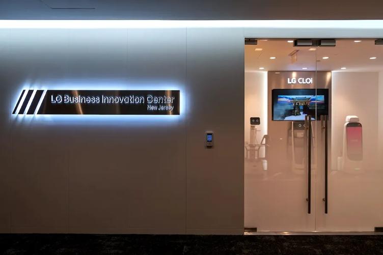 A backlit silver sign reading LG Business Innovation Center New Jersey besides a pair of sleek glass doors. Through the doors, the Business Innovation Center itself is visible, with a digital signage display and three CLOi robots able to be seen.