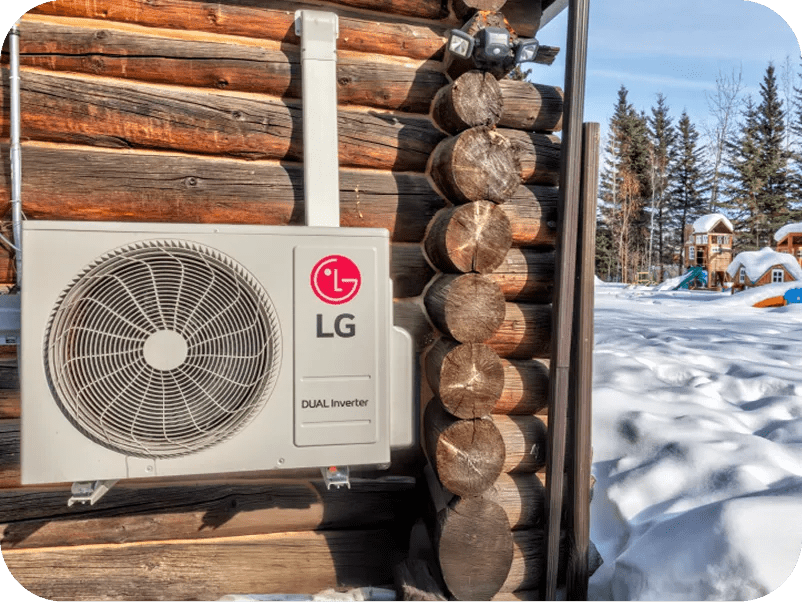 Outdoor LG Dual Inverter compressor unit mounted on the exterior wall of a log cabin, surrounded by snow, with a children's playground and forest visible in the snowy background.