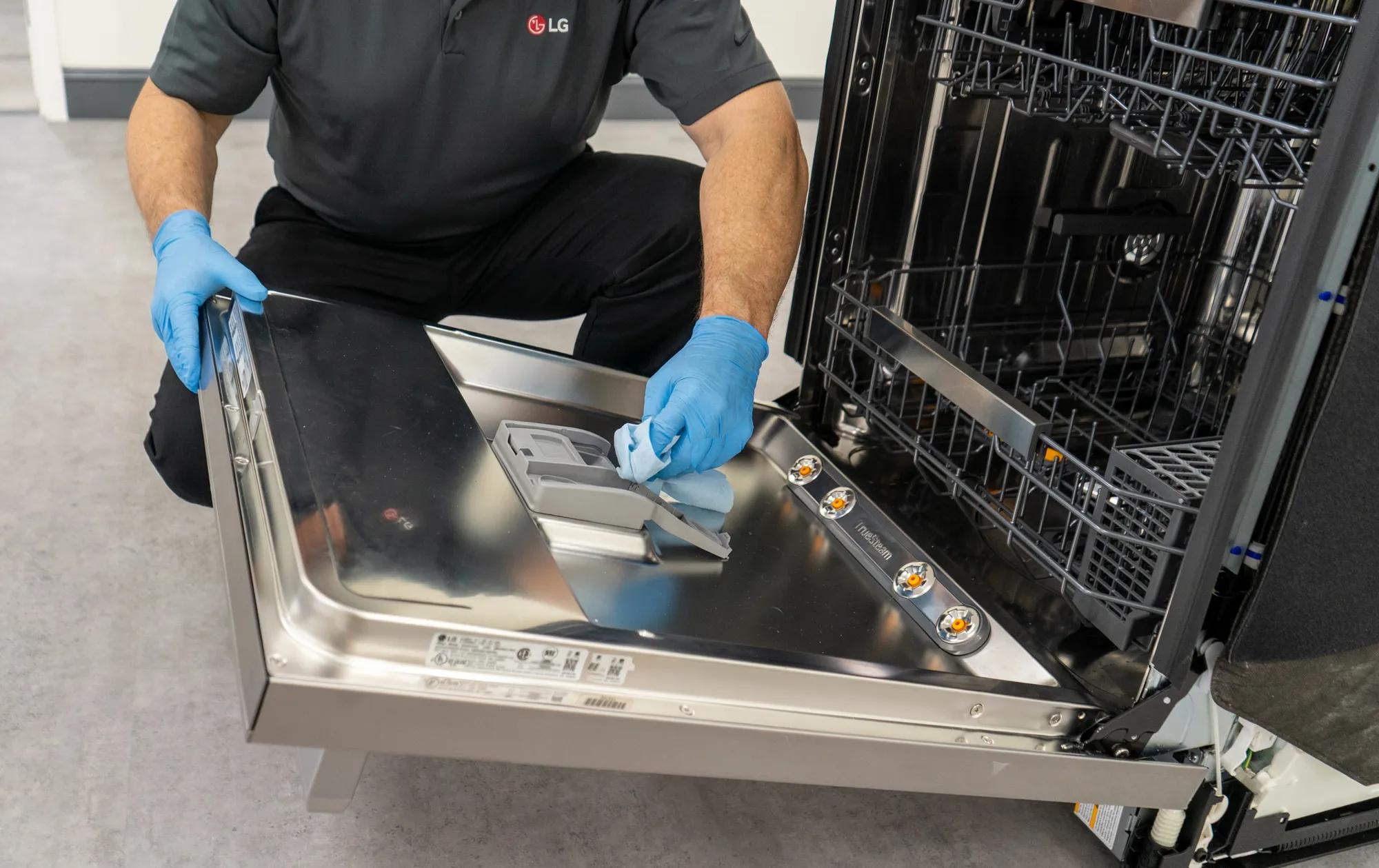An LG service expert wearing blue medical gloves using a cloth to sanitize the interior door and detergent dispenser of a LG dishwasher.