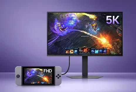 A handheld gaming device is connected to a large desktop monitor. Both screens display the same fantasy game scene featuring colorful magical effects and characters in combat. The monitor is significantly larger and shows the image in much higher detail than the handheld device. The setup sits on a smooth purple surface with a soft purple gradient background.