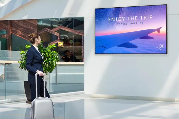 A flight attendant walking through an airport with suitcase. She's pausing to look at an LG Ultra HD display with a photo of an airplane's wing over sunset-colored clouds. The display says Enjoy The Trip, and below that, Travel Together.