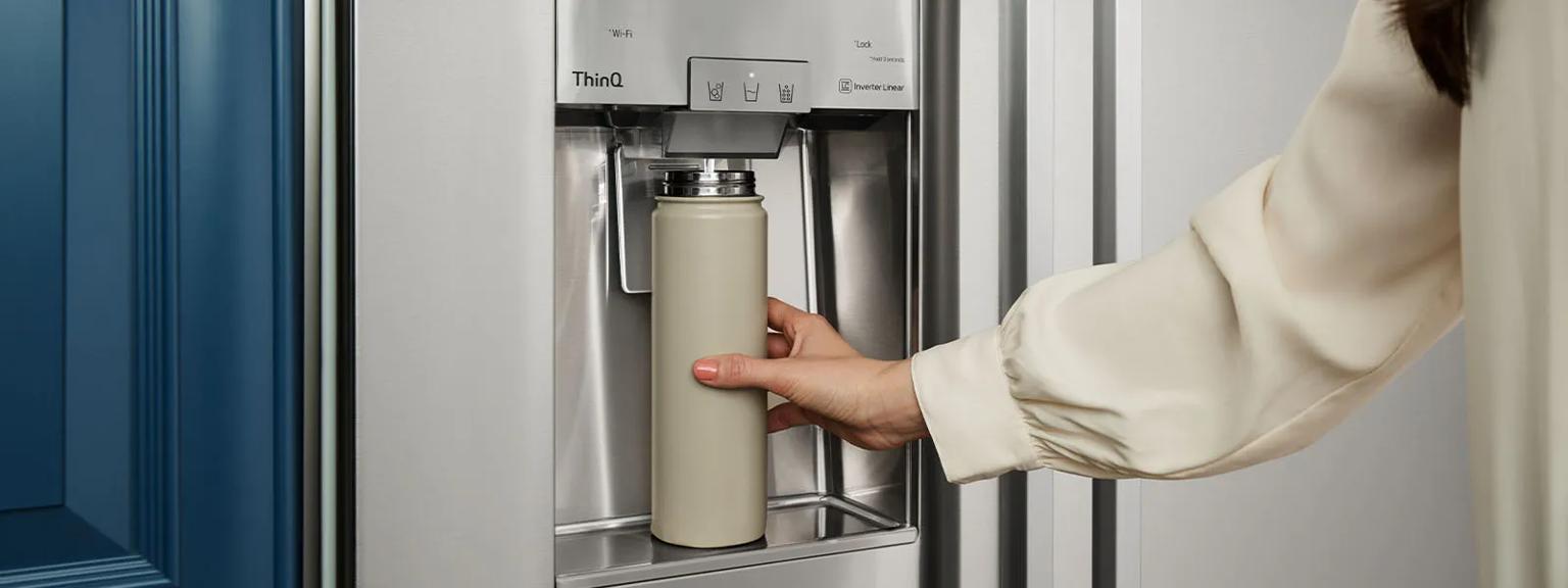 Hand holding refillable water bottle at an LG STUDIO Refrigerator water dispenser.