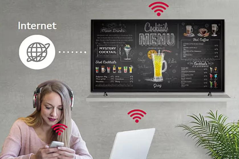 A woman with headphones using a smartphone and laptop, in front of a TV displaying a cocktail menu with Wi-Fi connectivity.