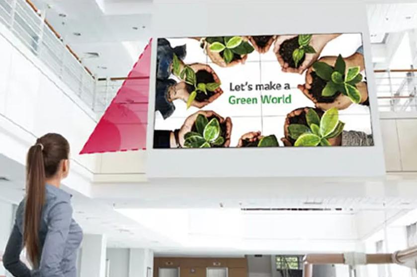 The image shows a woman with long brown hair, tied back, standing in a brightly lit, modern interior space. She is gazing up at a large digital screen mounted on a wall. The screen displays an image of several diverse hands holding small plants with vibrant green leaves, arranged in a circular pattern. The background of the screen is white, providing contrast to the greenery. Adjacent to the plants is text with an environmental message. The space around her features white walls, a high ceiling, and a mezzanine level with glass railings. A red triangular design element decorates the upper left portion of the wall near the screen.