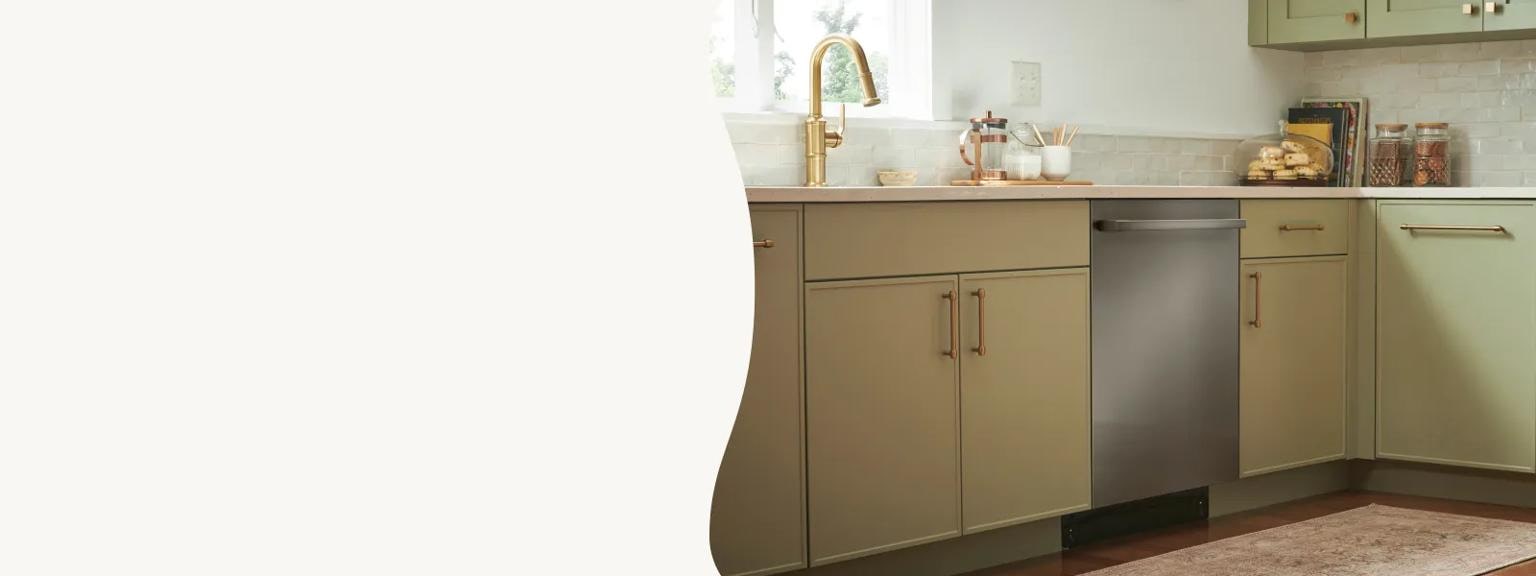 A sleek, stainless steel dishwasher is seamlessly integrated into sage green cabinetry with gold hardware. The warm kitchen space is accented by a gold faucet, a light-tiled backsplash, and a patterned runner rug on wood floors.