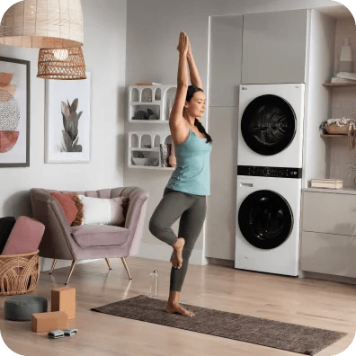 A woman practicing yoga in a tree pose on a mat in a modern living space, with a sleek white LG WashTower stacked washer-dryer unit integrated into the background cabinetry.