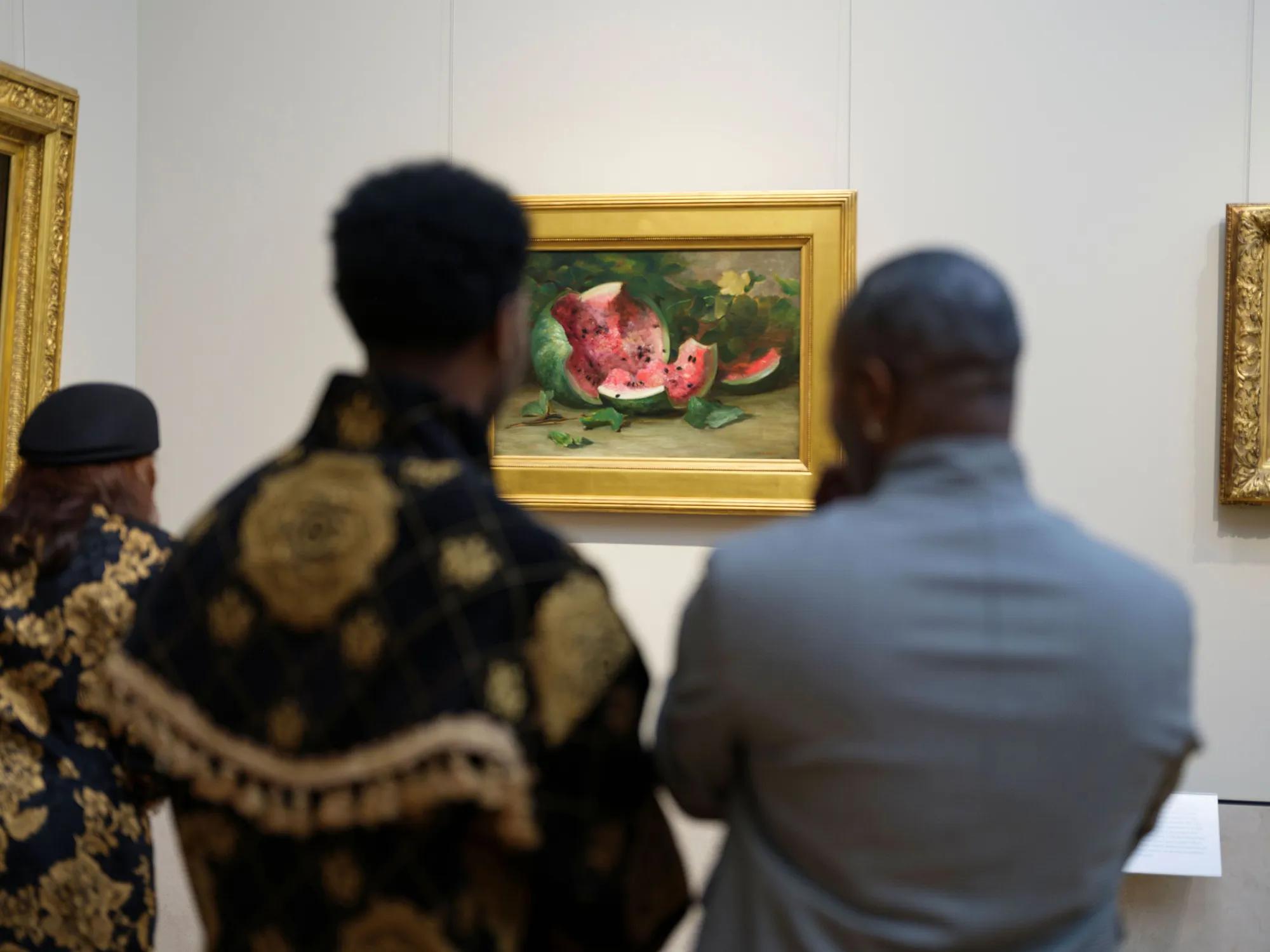 Three museum visitors, seen from behind and out of focus, look at a gold-framed still life painting of a broken-open watermelon on a gallery wall.