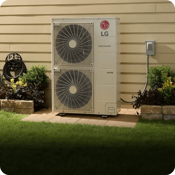 LG multi-zone outdoor HVAC unit with dual fans installed against the beige siding of a house, surrounded by flowers, and a coiled garden hose on a reel, resting on a stone-bordered patch of lawn.