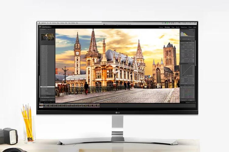 An LG 4K Ultra HD computer monitor sitting on a grey desk. Its display shows an editing software, with a main window displaying a photo of an ornate historical building. Also on the desk are some pencils in a cup and the edge of a camcorder otherwise out of frame.