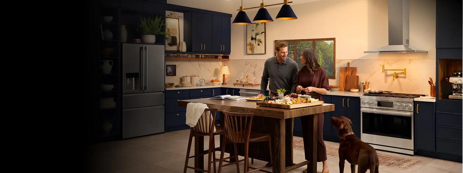 LG SIGNATURE Iconic Collection kitchen with refrigerator, dishwasher, range and wall-mounted range hood, in modern blue cabinetry, couple preparing food at island with dog nearby.