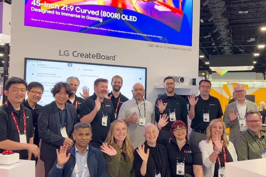 A large LG team posing in front of an LG CreateBoard at an education tradeshow, all waving at the camera.
