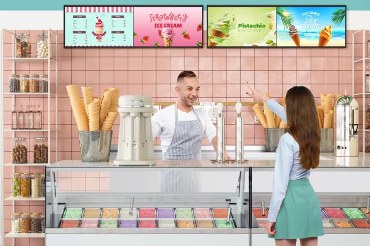 A girl at an ice cream shop's counter, with a cheerful employee waiting to take her order. She's pointing at the LG Ultra Stretch signage hanging behind the counter, which displays menu pricing as well as advertising different flavors.