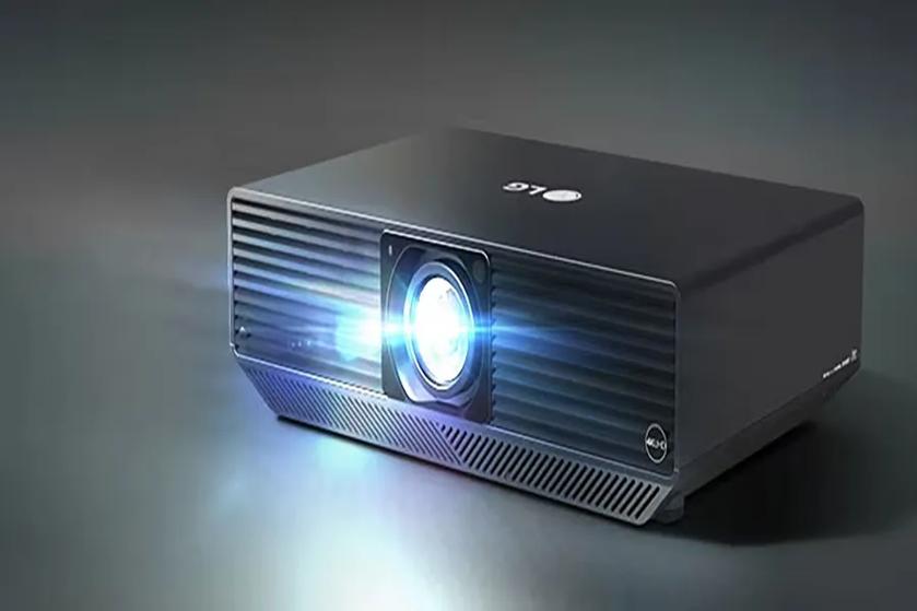 The image shows a sleek, dark gray projector centrally positioned on a smooth, gradient background that transitions from light to dark gray. The projector has sharp, rectangular edges and a modern design. Its front side features a prominent lens emitting a bright, blue-white light, creating a reflective glow on the surface below. The lens is surrounded by a ribbed section that likely aids in ventilation. On the top of the projector, there is a brand logo in white. The overall design is minimalist and professional, suitable for both office and entertainment settings.