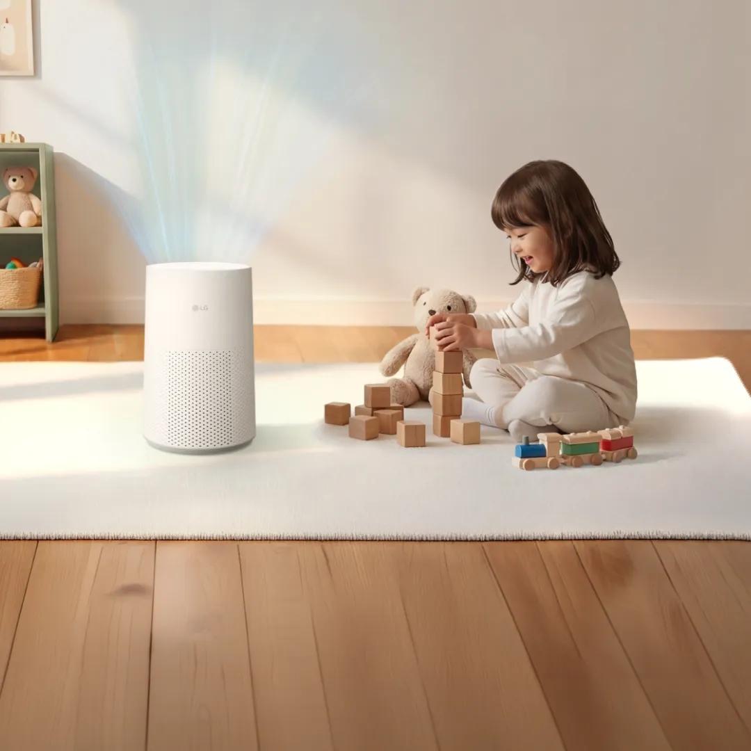 A young girl smiles while stacking wooden blocks on a white rug, with a teddy bear and toy train beside her, as a white LG air purifier releases a visible stream of clean air nearby in a sunlit room with wood flooring and a small bookshelf of toys.