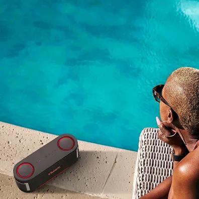 Young woman wearing sunglasses sits poolside next to an LG xboom portable speaker.
