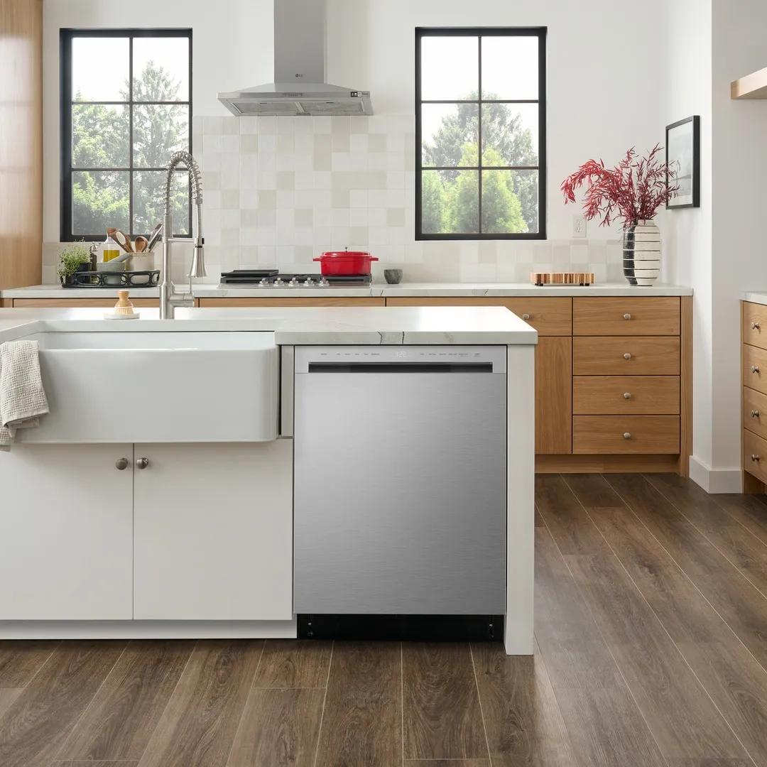Kitchen photo: a stainless steel LG dishwasher built into a white island with a farmhouse sink, beside a wood-cabinet kitchen featuring a stainless range hood, cooktop, and windows onto greenery.