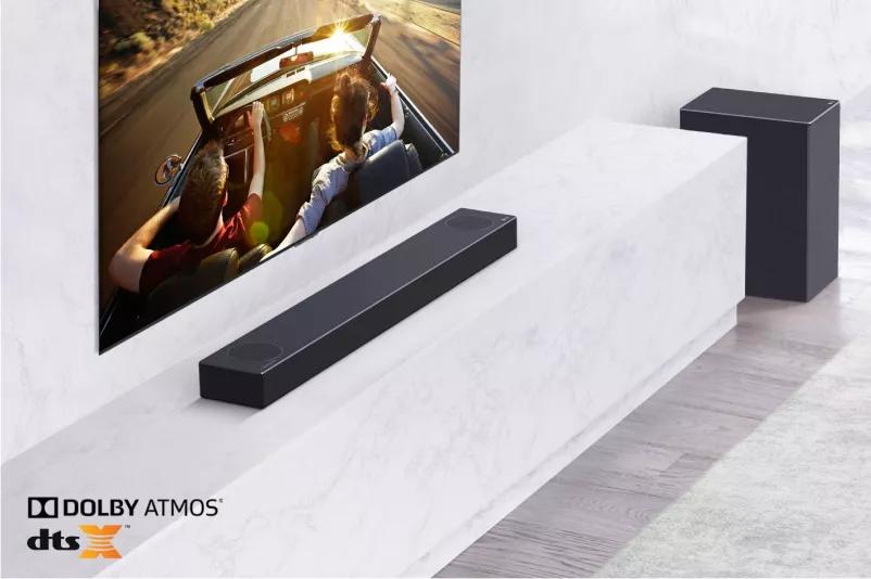 TV is on the wall LG Soundbar is below on a white marble shelf with a sub-woofer to the right TV shows a couple in a car