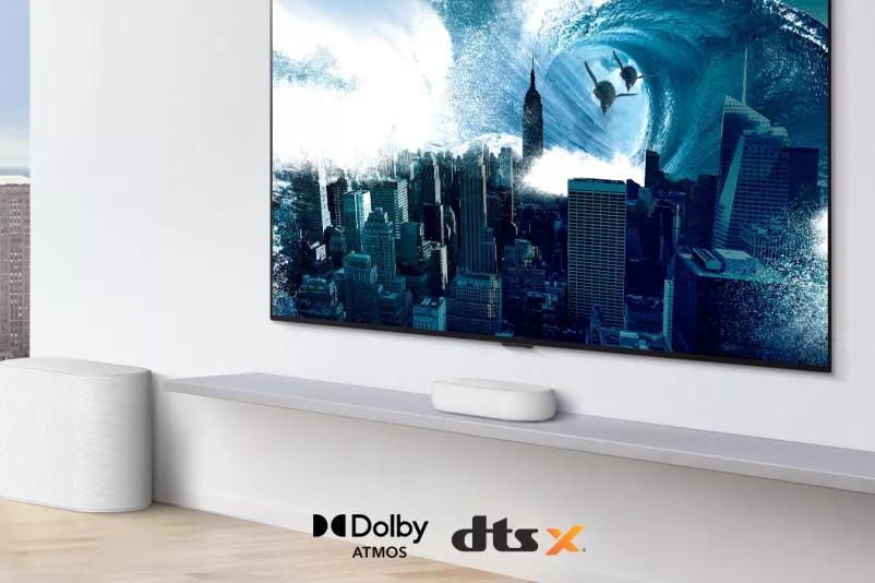 TV is on the wall TV shows a big swirling wave hitting the entire city LG Eclair is right below TV on a white shelf with a sub-woofer right next Dolby Atmos and DTS X logo shown on middle bottom of image