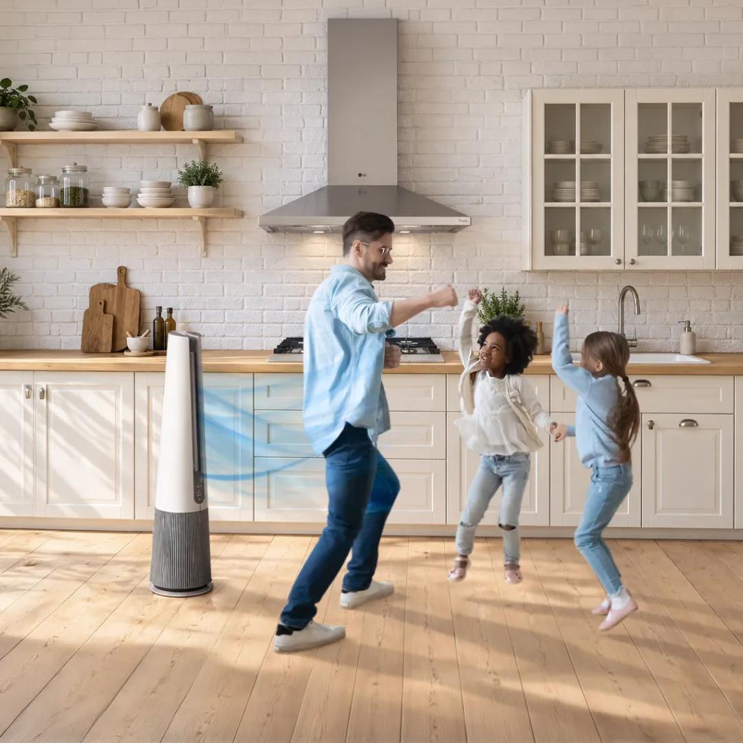 A man and two girls dance happily in a bright kitchen while a tall tower air purifier fan blows cool air nearby.