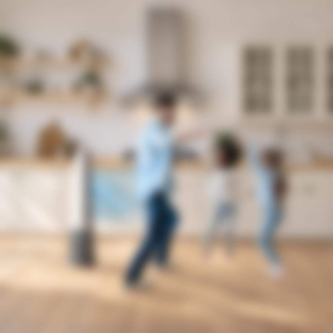 A man and two girls dance happily in a bright kitchen while a tall tower air purifier fan blows cool air nearby.