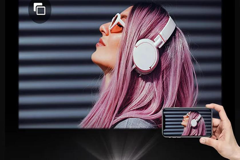 The image showcases a person with long pink hair and wearing white headphones and white-framed sunglasses. They are positioned in profile, facing left, against a backdrop of horizontal black stripes. The foreground features a hand holding a smartphone, which displays a smaller version of the same image of the person. The composition highlights the vivid pink hair and white accessories, creating a striking contrast with the dark background.