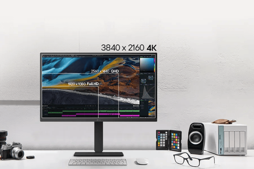 Front view of a white LG monitor on a desk, displaying layered screen resolutions for comparison, with cameras, books, color chips, and speakers arranged around it.