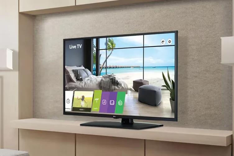 A modern hotel room with an LG Commercial Lite Hotel TV. The TV shows a relaxing beach scene with UI controls overlaid at the bottom.