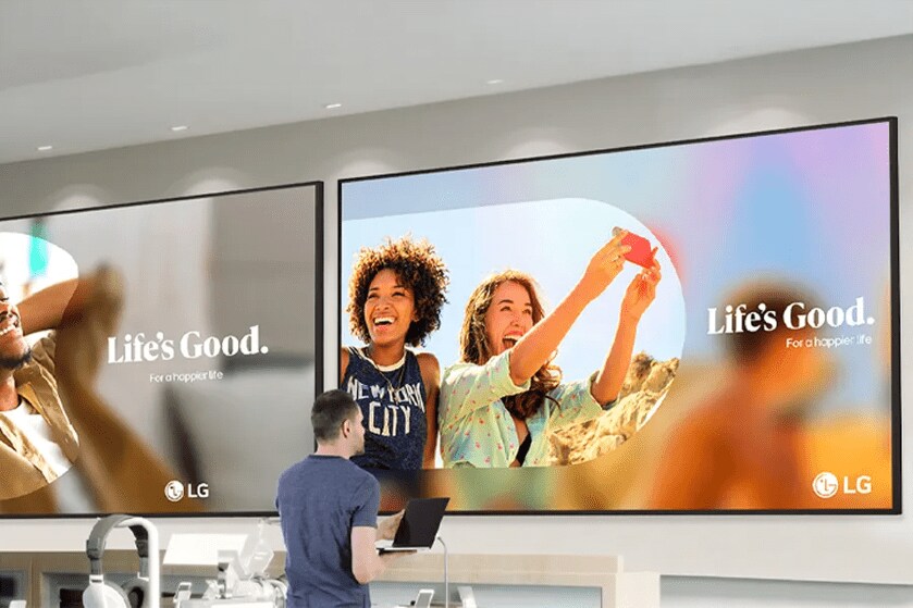 A person with a laptop standing in front of two large LG screens displaying promotional content with text "Life's Good. For a happier life."