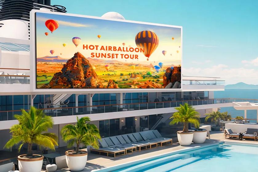 A large LED display is mounted on the exterior wall of the superstructure facing the pool area on the Lido Deck of a luxury cruise ship, clearly and brightly showing advertisements on a large screen.