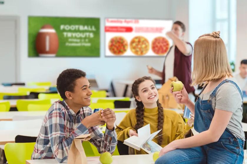 Teens sit in a cafeteria, smiling and talking. Behind them, LG Displays advertise the pizza lunch options and upcoming football tryouts.