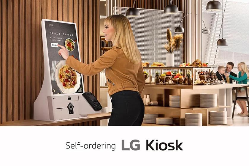 The image shows a modern restaurant interior with a focus on a self-ordering kiosk. A woman with long blonde hair, wearing a tan jacket and black pants, interacts with the touchscreen of the kiosk. The kiosk features a sleek white base with a card reader attached. On the screen, there is an image of a pizza, and an "Order" button is displayed prominently. The background reveals a well-lit dining area with pendant lights, neatly arranged food displays, and shelving stacked with plates. A family is visible seated at a table enjoying their meal.