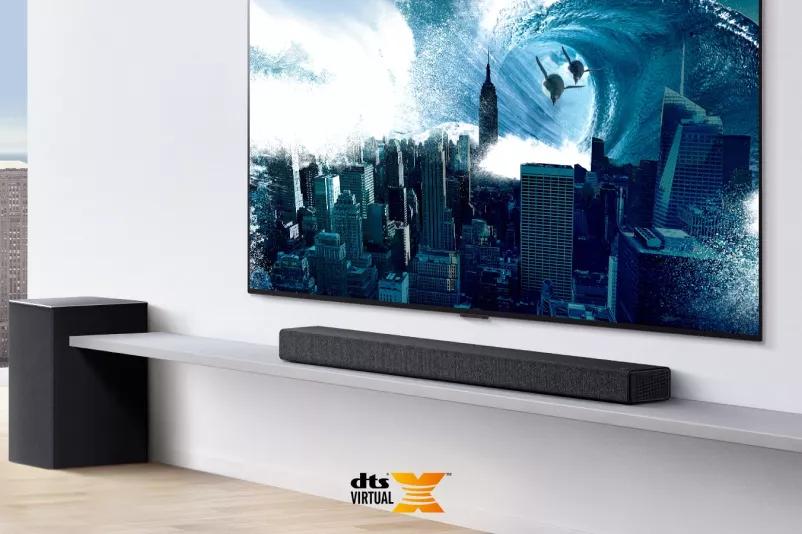 TV is on the wall TV shows a big swirling wave hitting the entire city LG Soundbar is right below TV on a white shelf with a sub-woofer right next Dts Virtual X logo shown on middle bottom of image