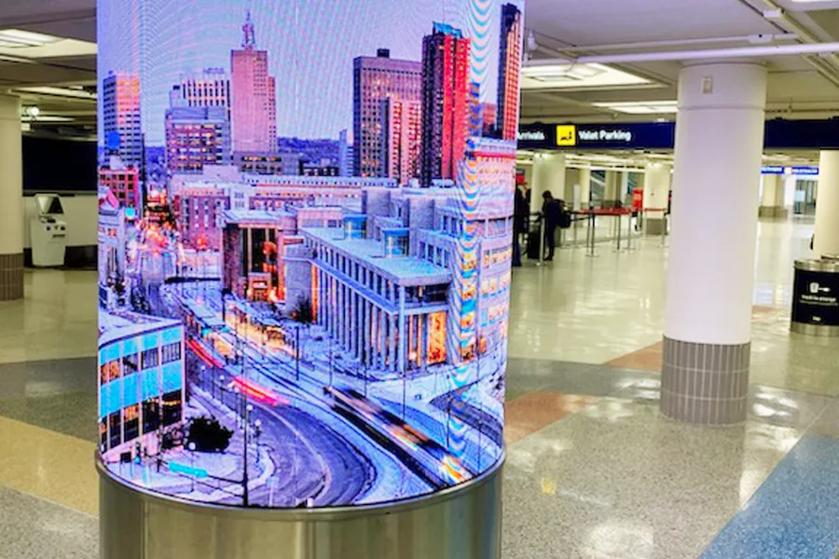 Curved LG OLED signage wraps around a column inside an airport terminal, displaying a brightly lit city scene.