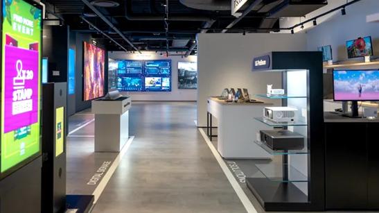 Interior of the LG Business Innovation Center in New Jersey. The photo shows a walkway formed by various brightly lit signage displays on the left, and display tables of monitors, laptops, and projectors on the right. At the far end of the space, large scale DVLED displays are visible.
