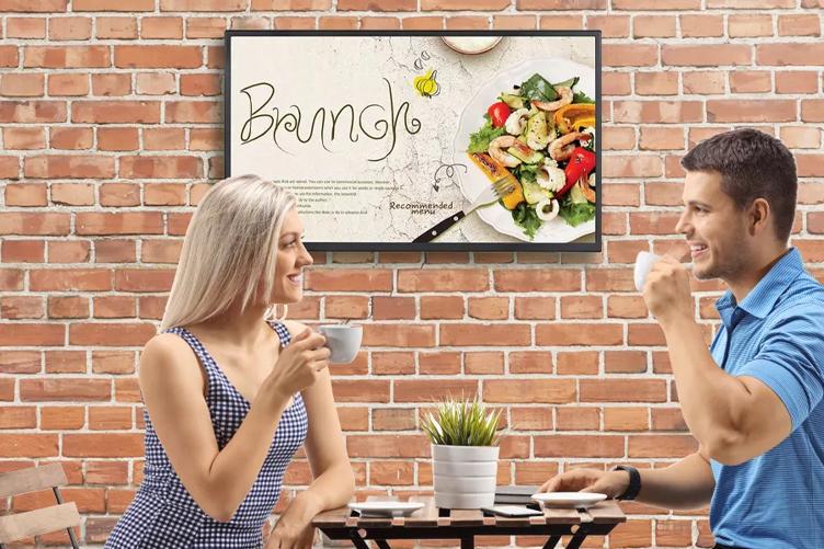 A couple drink coffee at a cafe style table. A brick wall is behind them, and an LG Full HD signage hangs on it advertising a Brunch menu.