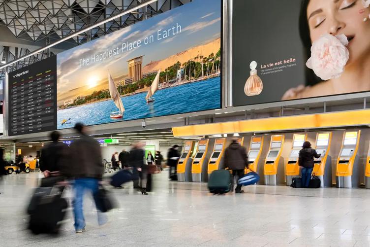 Busy airport terminal with travelers, two large ads, a departures board, and check-in kiosks.