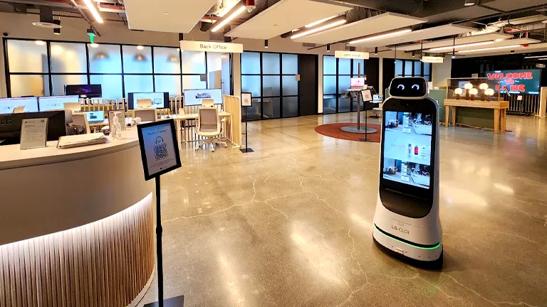 Interior of the LG Business Innovation Center in Los Angeles, with a check in desk to the left and a CLOi GuideBot to the right. Beyond these is an open space with tables displaying computer monitors, an LG gram laptop lounge area, and digital signage.