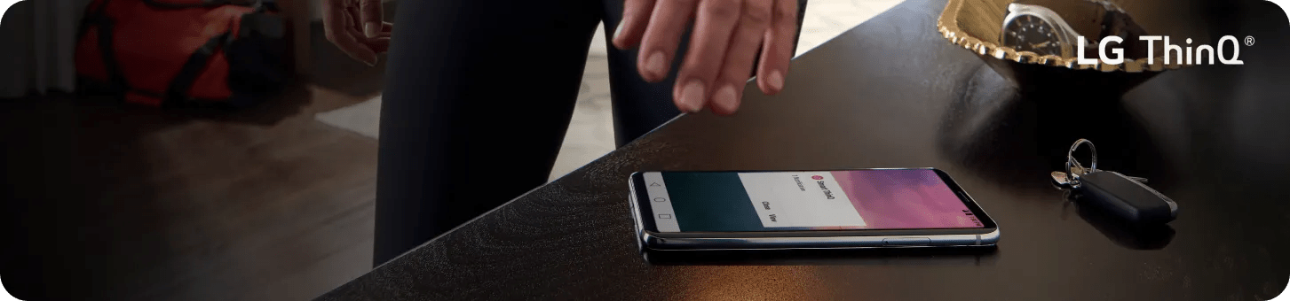 Smartphone resting on a dark tabletop displays an LG ThinQ app screen, with a hand nearby and keys placed beside it in a home setting. In the corner is a badge that says, 'Brought to you by America's most reliable line of appliances.'