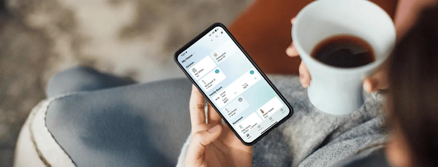 A person holds a smartphone with the LG ThinQ app open, managing smart home devices while drinking coffee.