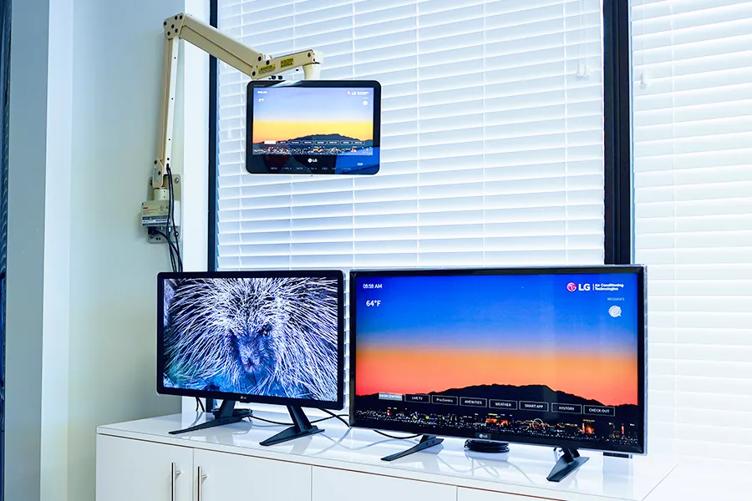 Two hospitality TVs stand on a display shelf at the LG Atlanta Business Innovation Center, showing bright wallpaper images of a bird and a mountain landscape. Above them, a smaller hospital monitor for healthcare patients is mounted on a hospital TV arm, with a natural mountain scene as the background of its UI. 