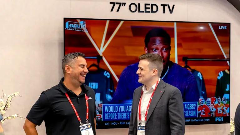 Two LG employees stand in front of a 77" LG OLED Hospitality TV at HITEC 2025, smiling as they chat. The TV is showing a close-up of a sports commentator.