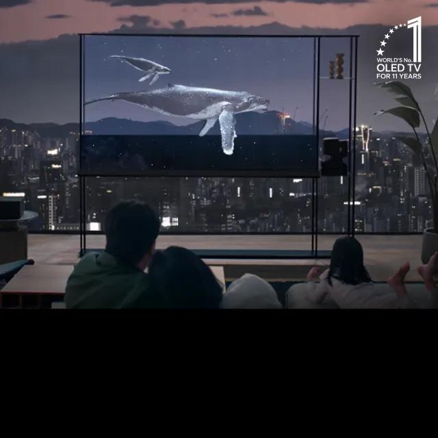 An image showcasing a modern, transparent OLED TV in a cozy living room with a cityscape in the background. The TV displays an underwater scene with a humpback whale and its calf swimming, creating a vivid and immersive viewing experience. A family is seated on a couch, watching the screen in awe, with the text overlay 'World's No. 1 OLED TV for 11 Years' in the top-right corner.