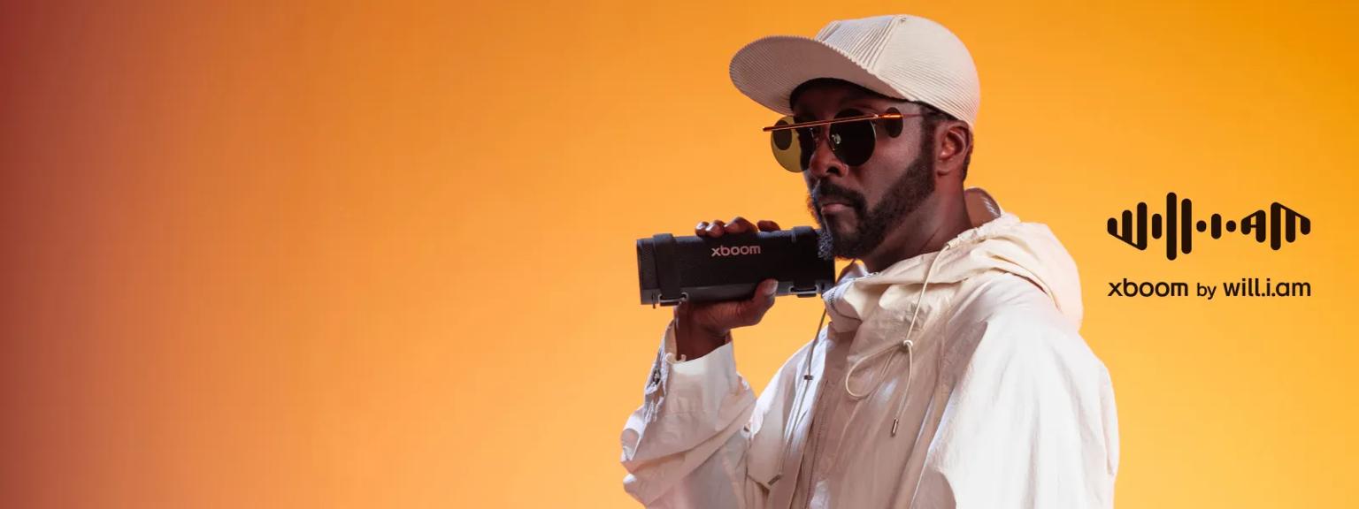 Music artist will.i.am holds an xboom speaker in before an orange background. Besides the image appears text reading “xboom by will.i.am”.