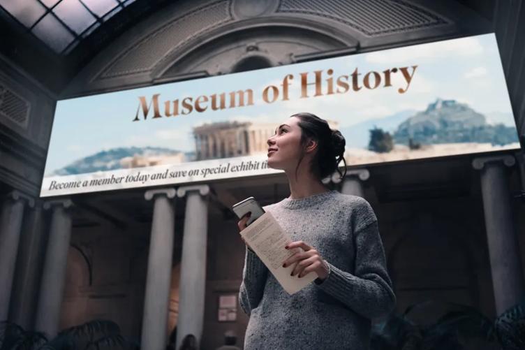 A woman walks into an elegant museum foyer. Large direct view LED on the wall above her reads Museum of History with an image of ancient Greek ruins. 