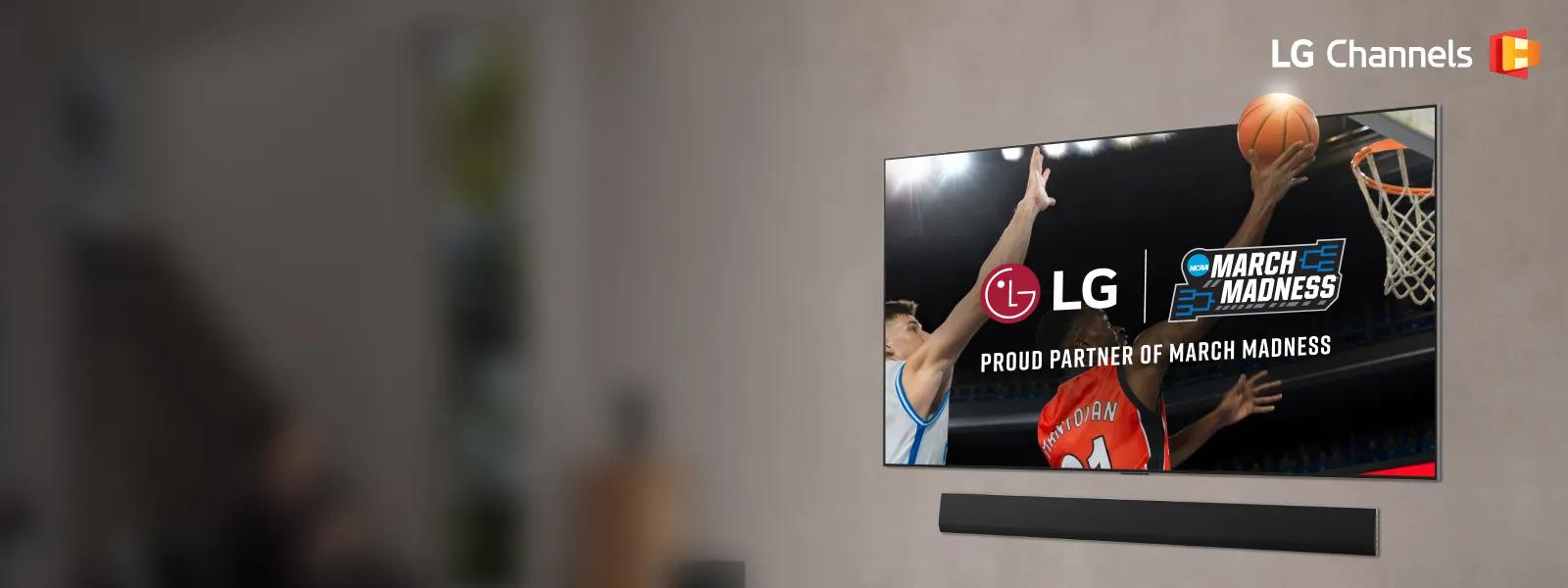 An LG TV mounted on a wall displays a close-up of a basketball player in a red jersey performing a layup being challenged by a player in a white jersey as a fan cheers on in a modern living room. Partnership lockup of LG and NCAA March Madness is overlayed on the TV. LG Channels logo.