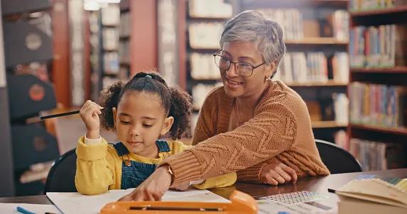 A grandmother helping a child learn, teaching and writing in education, language in a school library.
